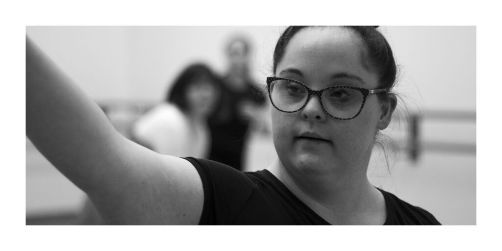 A black and white headshot of a young woman, Emily. Her hair is dark coloured and tied up. She is wearing a pair of dark rimmed glasses and a short sleeved dark coloured tee shirt. Her gaze is towards the left, following the direction of her outstretched arm.  There are two other people in the background, but they are out of focus and unidentifiable.