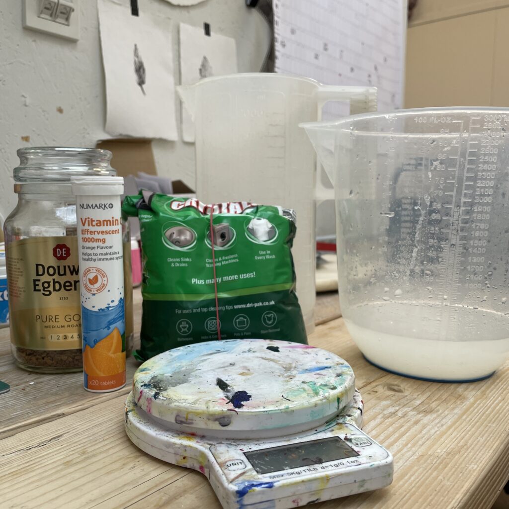 A wooden desk with everything you need to make caffenol-c developer. A set of digital scales, a couple of plastic jugs, a bag of soda crystals, a tube of vitamin c and a jar of instant coffee.