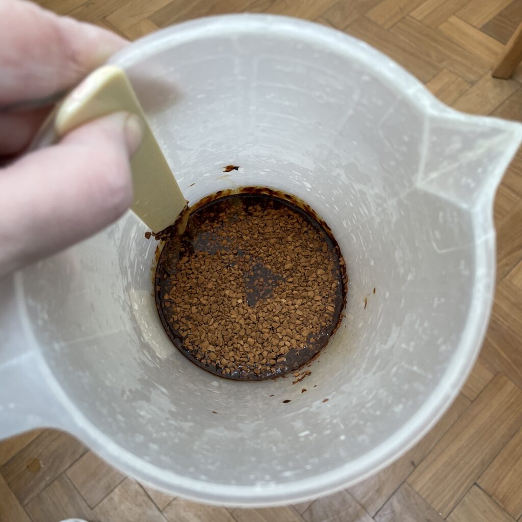 An overhead view of coffee granules dissolving in a jug of liquid, a hand is holding a stirrer in the left hand side of the jug.