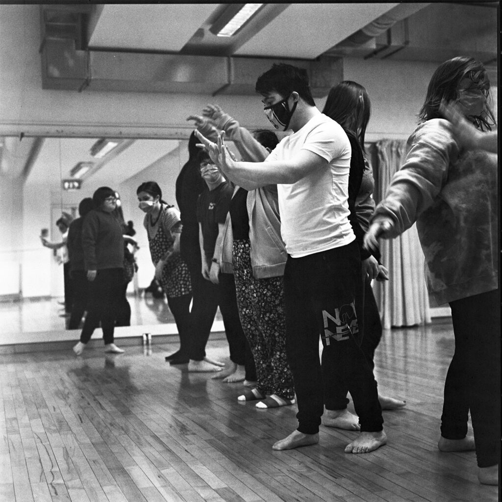 A black and white image of a group of dancers stood in a line. Several of the dancers are making shapes with their arms.
