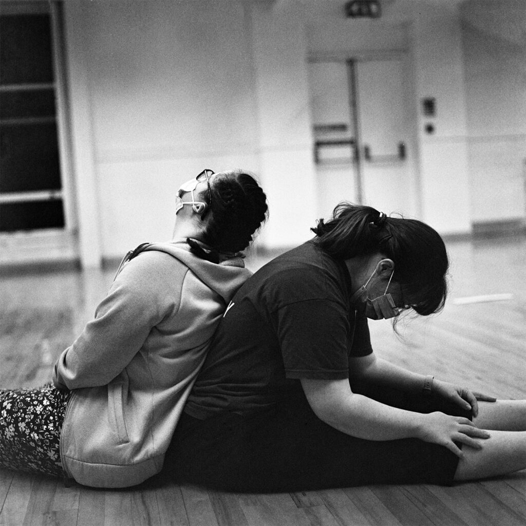 Two dancers sat back to back on the wooden studio floor. The image is black and white.