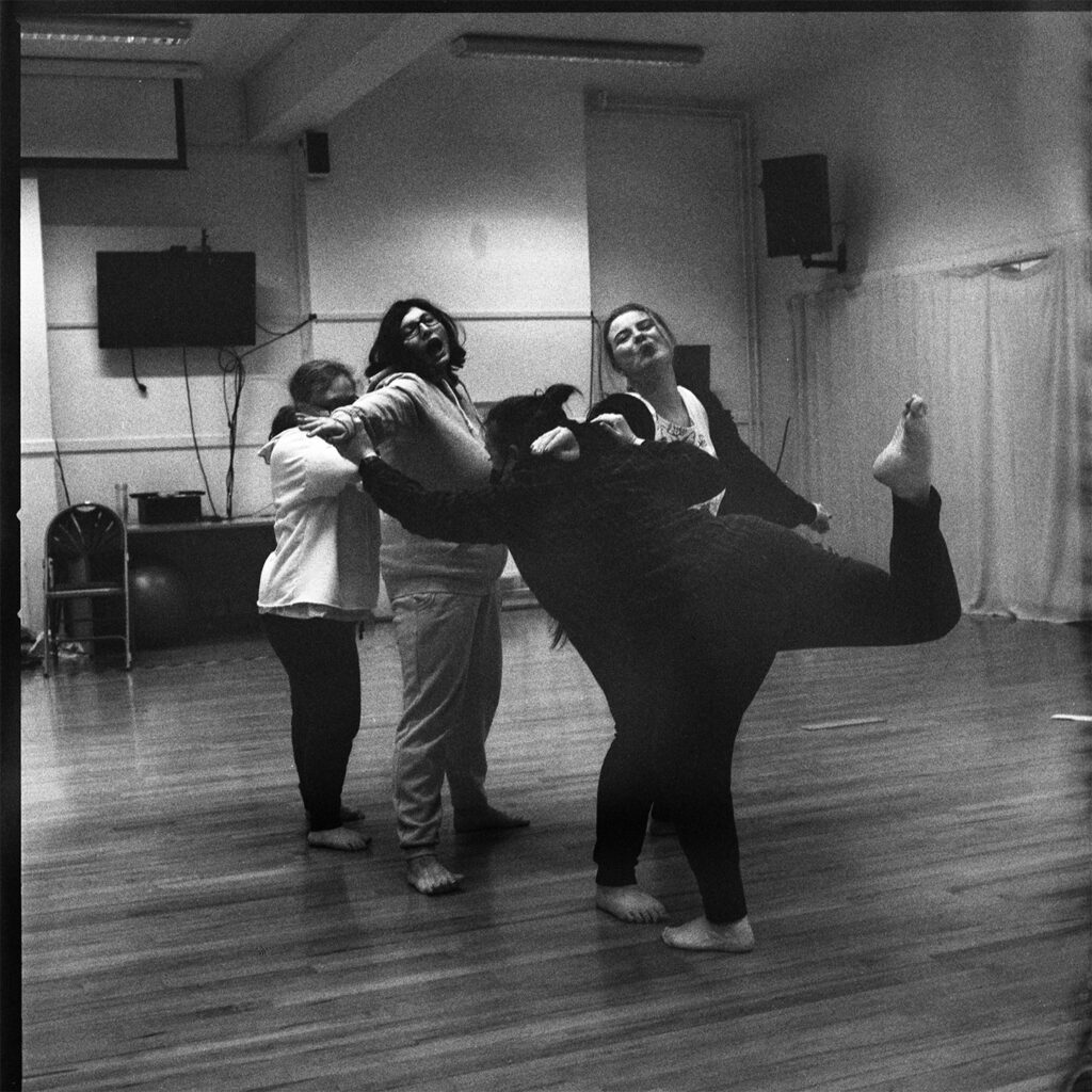 A group shot of four dancers. At front of the group one of the dancers is balanced on one leg, stretching in front of the other three.