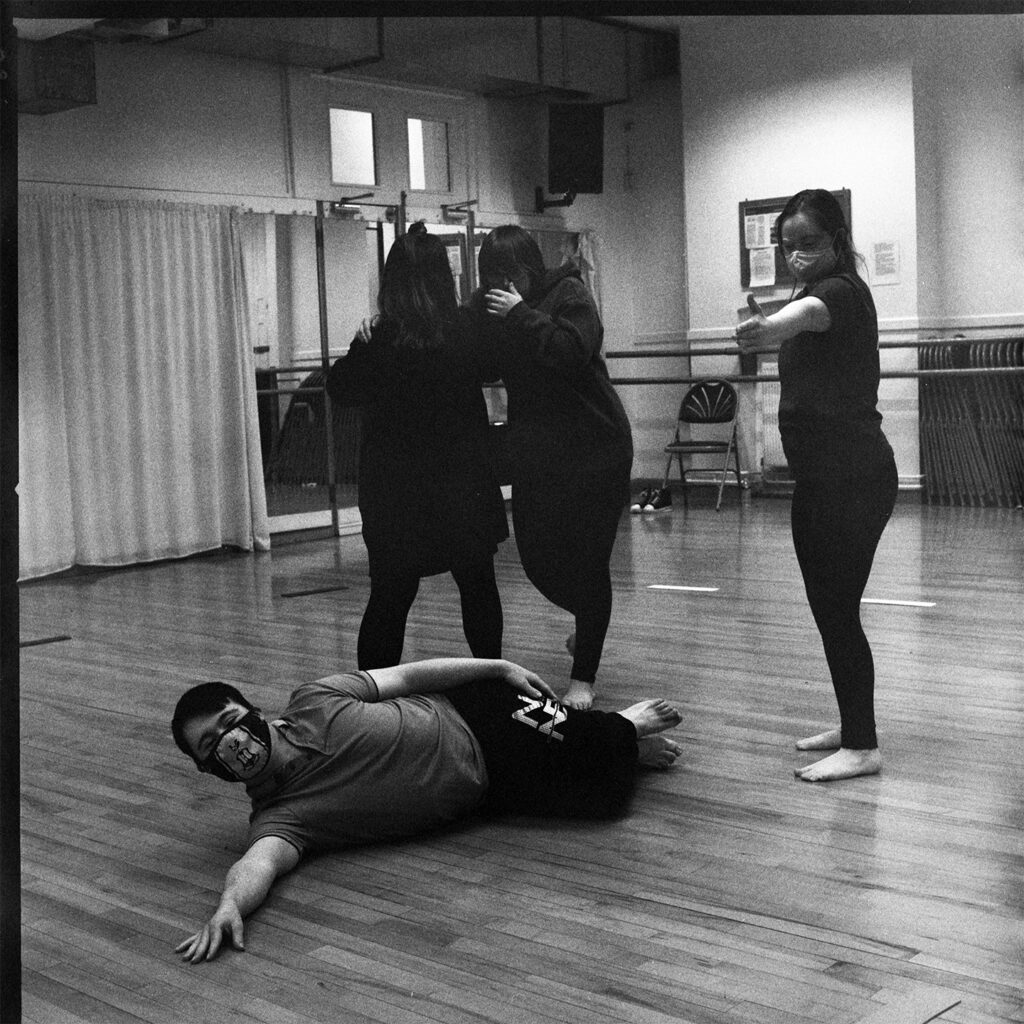 A group shot of four dancers. One of the dancers is posing, laying on the flour. The other three are stood around them.