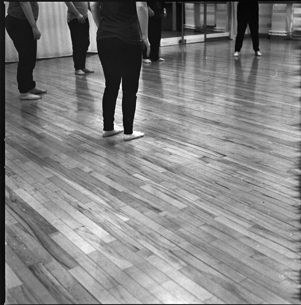 A black and white photo of dancers stood in their rehearsal studio. The dancers are only visible from the waist down