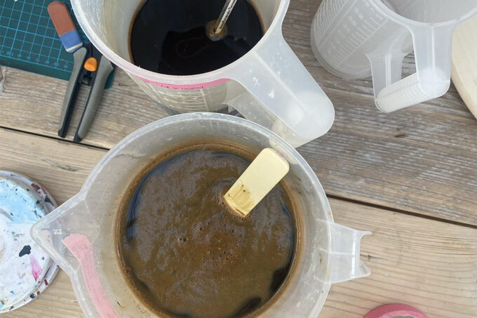 An overhead view of two jugs of caffenol-c developer on an untidy desk. One jug has a plastic stirrer in it and the other has a thermometer in it.