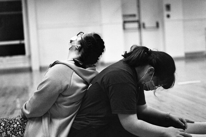 Two dancers sat back to back on the wooden studio floor. The image is black and white.