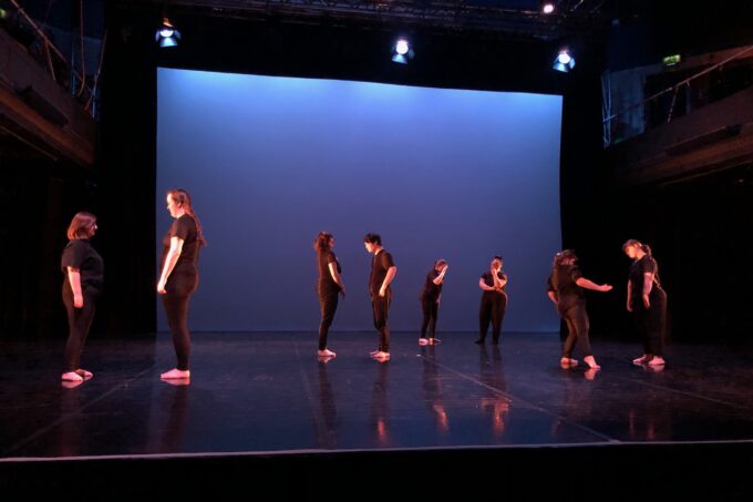 Seven dancers are spread out across a wide stage, standing in pairs facing one another. They are caught in various movements. The dancers are all barefoot and wearing black. Behind them is a large screen and above are three stage lights. The area around them is dark.